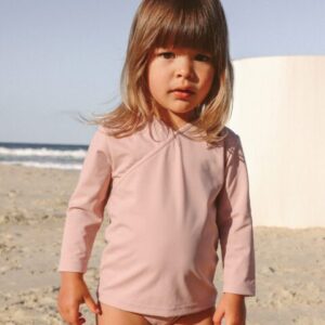 A young child in a pink Ada Rash Shirt - Rose Colour stands on a sandy beach with the ocean in the background.