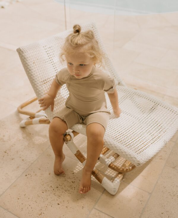 A toddler in a Zimmi Onesie - Sand with a topknot hairstyle sitting thoughtfully on a white poolside lounge chair.