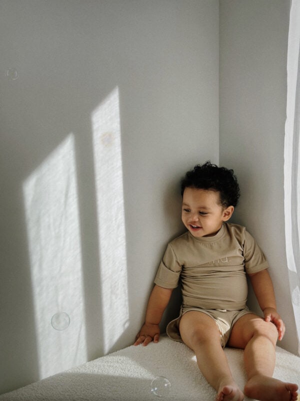 A young child sitting on a bed, smiling while looking at floating bubbles in a softly lit room with sunlight casting shadows on the wall, wearing an Ina Rash Shirt - Sand Colour.