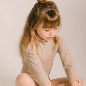 A little girl sitting on the floor in a tan June Long Sleeve One-Piece.