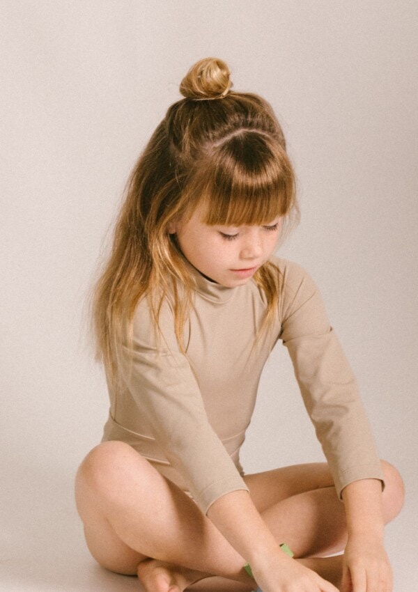A little girl sitting on the floor in a tan June Long Sleeve One-Piece.