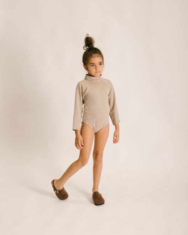 A little girl wearing a June Long Sleeve One-Piece bodysuit and brown slippers.