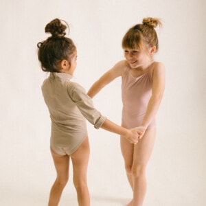 Two little girls are holding hands while wearing the June Long Sleeve One-Piece in a studio.