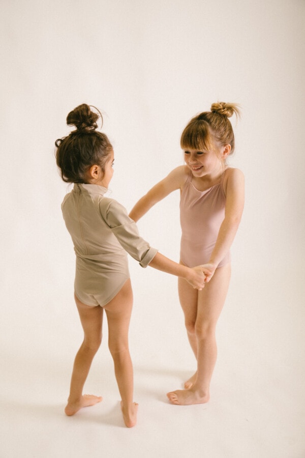 Two little girls are holding hands while wearing the June Long Sleeve One-Piece in a studio.