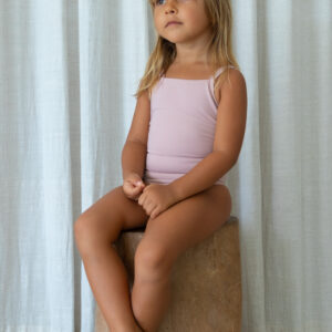 A little girl in a pink swimsuit, the Mara One-Piece, sitting on a wooden stump.