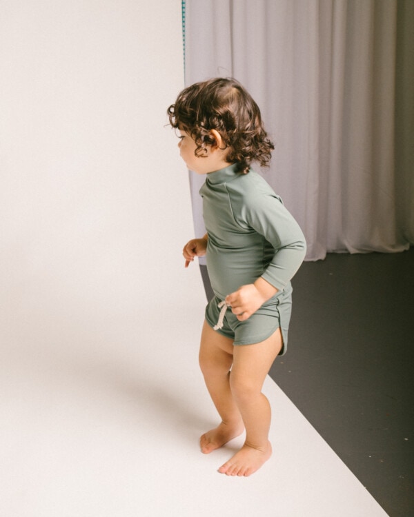 A baby standing on a white background in Mesa Trunks.