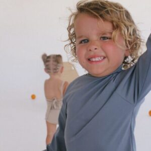 A smiling child with curly hair poses playfully in front of a white background, wearing the Nella Rash Shirt - Mineral Colour, with another child visible in the background.