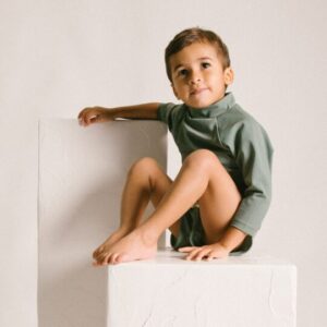 A young child sitting on a Nella Rash Shirt - Moss Colour against a neutral background.