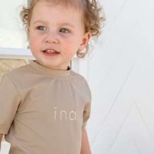 A little girl wearing a Essentials Range - Ina Rash Shirt - Sand Colour with the word ina on it.