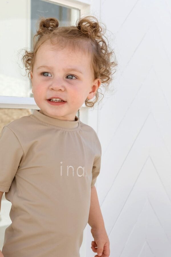A little girl wearing a Essentials Range - Ina Rash Shirt - Sand Colour with the word ina on it.