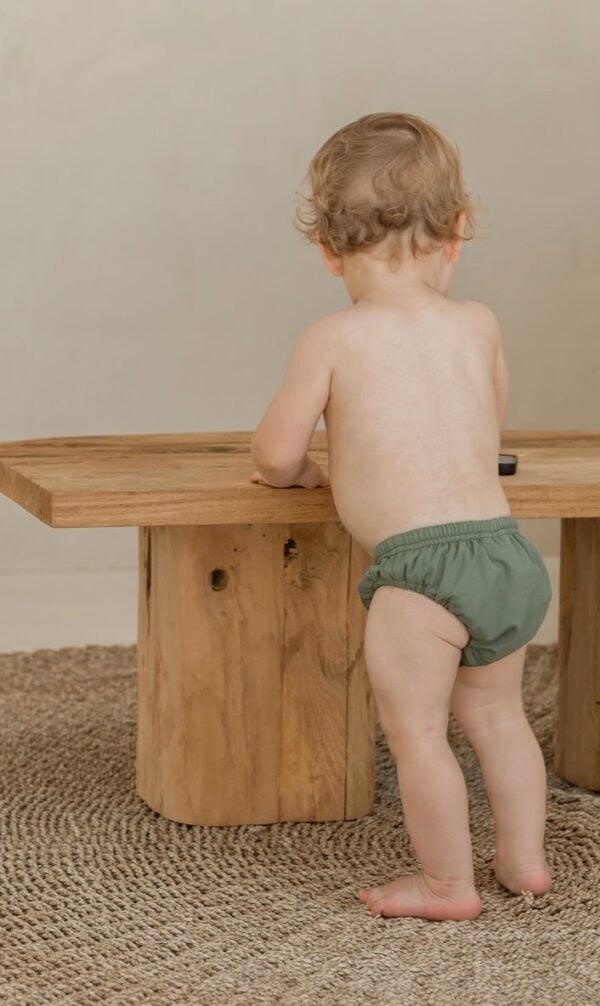 A baby in a Essentials Range - Lumi Brief Swim Nappy - Moss Colour standing next to a wooden table.