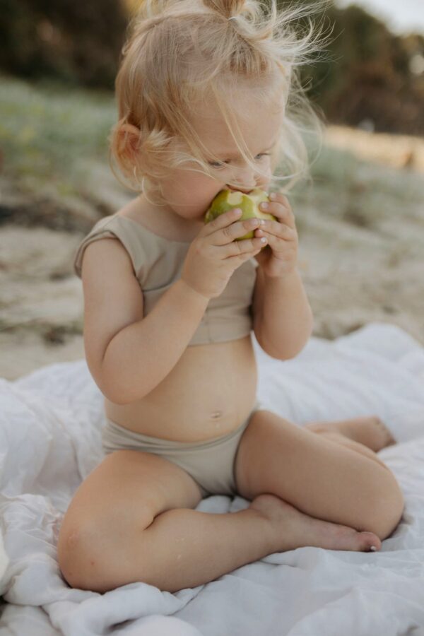A little girl eating an Essentials Range - Arla Bikini - Sand Colour on the beach.