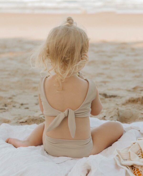 A little girl sitting on the beach in the Essentials Range - Arla Bikini - Sand Colour.