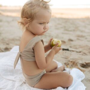 A little girl sitting on the beach wearing the Essentials Range - Arla Bikini - Sand Colour.