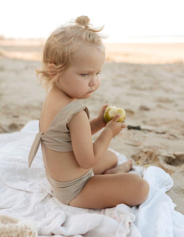 A little girl sitting on the beach wearing the Essentials Range - Arla Bikini - Sand Colour.