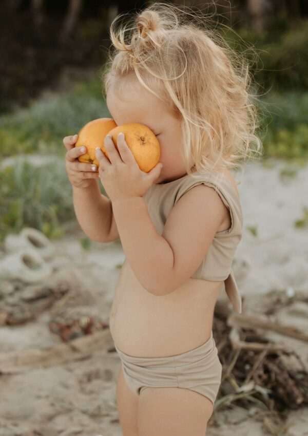 A little girl eating an Essentials Range - Arla Bikini - Sand Colour on the beach.