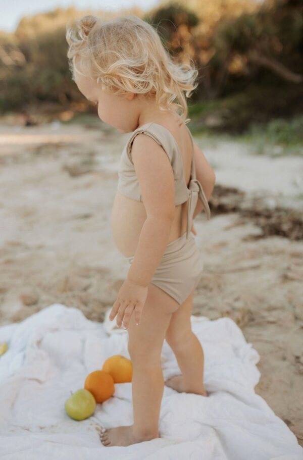 A little girl standing on the beach in the Essentials Range - Arla Bikini - Sand Colour.