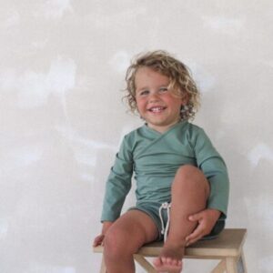 A young child with curly hair smiling while sitting on a wooden stool against a plain background is wearing an Ada Rash Shirt in Moss Colour.