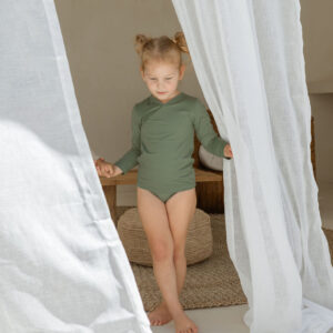 A little girl in a green bodysuit standing under the Essentials Range - Ada Rash Shirt - Moss Colour