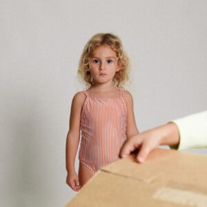 A little girl standing next to the Playtime Collection - Mara One-Piece - Mandarine Stripe.