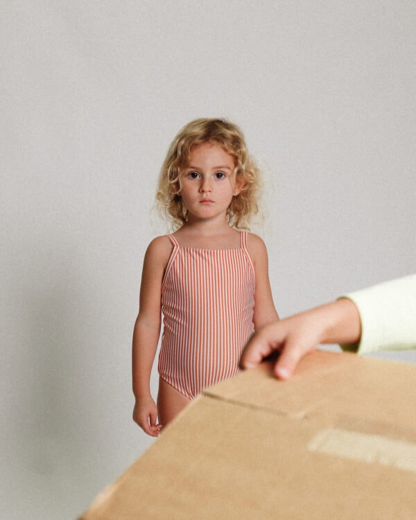 A little girl standing next to the Playtime Collection - Mara One-Piece - Mandarine Stripe.