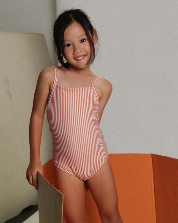 A little girl in the Playtime Collection - Mara One-Piece - Mandarine Stripe swimsuit standing on top of an orange box.