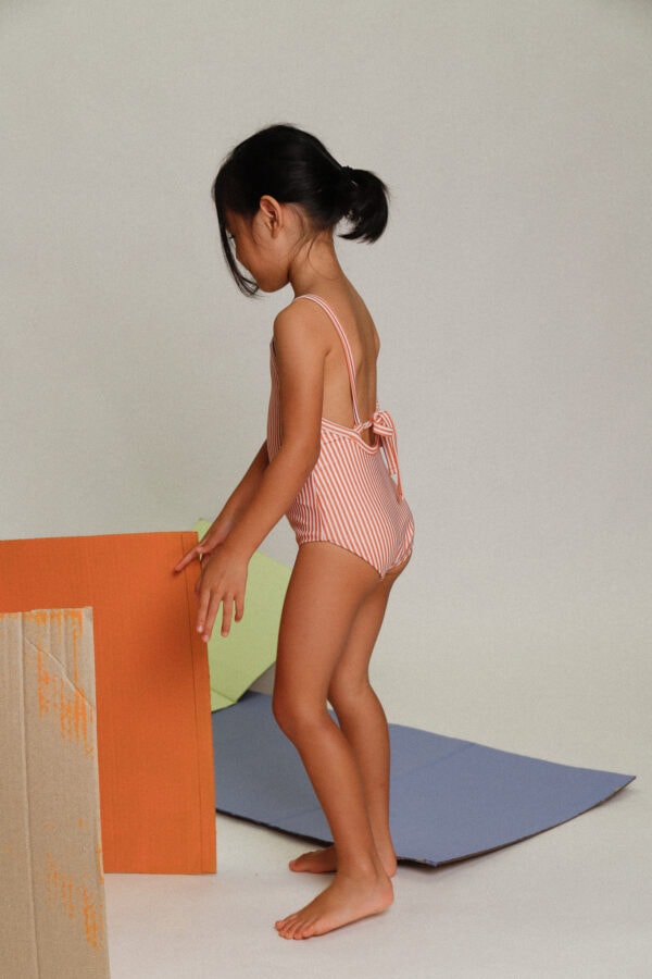 A little girl in a Playtime Collection - Mara One-Piece - Mandarine Stripe swimsuit standing next to a cardboard box.