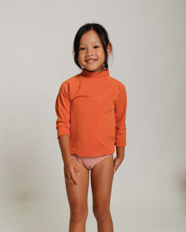 A little girl wearing the Playtime Collection - Nella Rash Shirt in Mandarin and striped shorts.