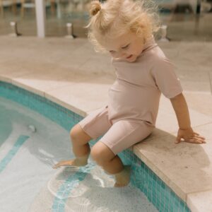A young child with blond hair wearing a Zimmi Onesie - Rose sits by the poolside, dipping a foot in the water.