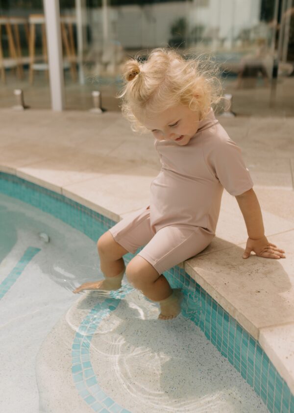 A young child with blond hair wearing a Zimmi Onesie - Rose sits by the poolside, dipping a foot in the water.
