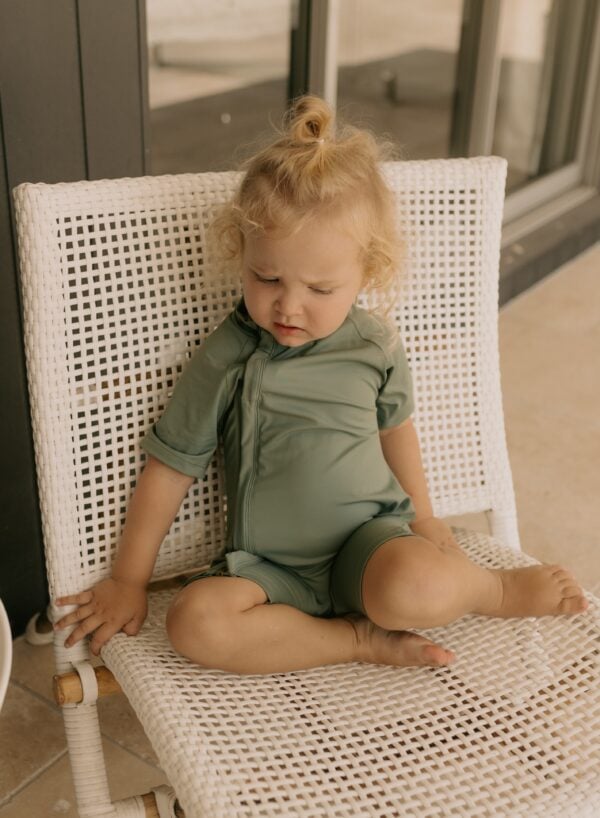 Young child in a Zimmi Onesie - Moss sitting contemplatively on a white wicker chair.