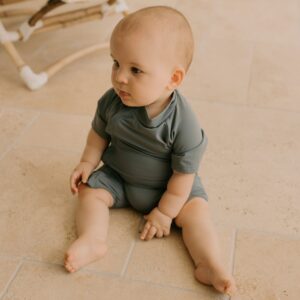 A baby sitting on a tiled floor, looking to the side, wearing a Zimmi Onesie - Mineral.