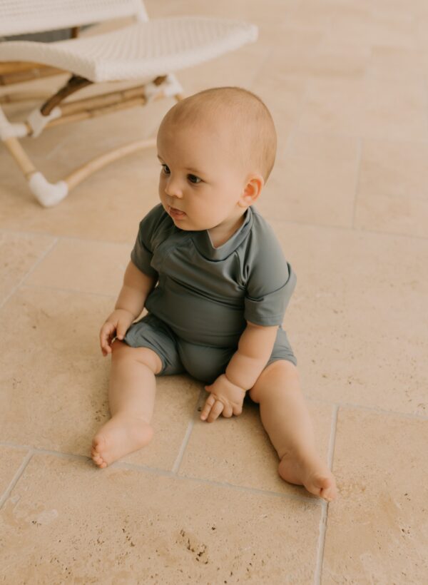 A baby sitting on a tiled floor, looking to the side, wearing a Zimmi Onesie - Mineral.