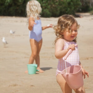 A little girl in an Azure & Apricot Gingham - Mara One-Piece swimsuit on the beach.