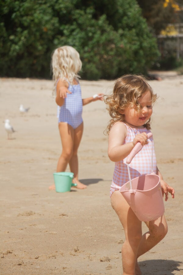 A little girl in an Azure & Apricot Gingham - Mara One-Piece swimsuit on the beach.