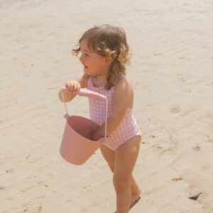 A little girl holding an Azure & Apricot Gingham - Mara One-Piece on the beach.