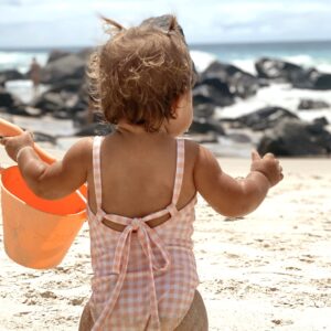A baby in a Mara One-Piece - Apricot Gingham swimsuit holding an orange bucket on the beach.