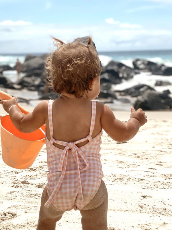 A baby in a Mara One-Piece - Apricot Gingham swimsuit holding an orange bucket on the beach.
