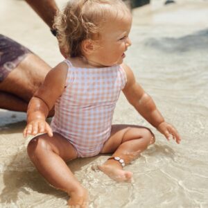 A baby girl wearing the Mara One-Piece - Apricot Gingham swimsuit on the beach.