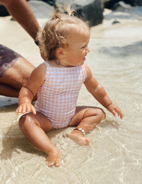 A baby girl wearing the Mara One-Piece - Apricot Gingham swimsuit on the beach.