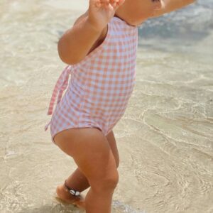 A baby girl in a Mara One-Piece - Apricot Gingham swimsuit standing in the water.