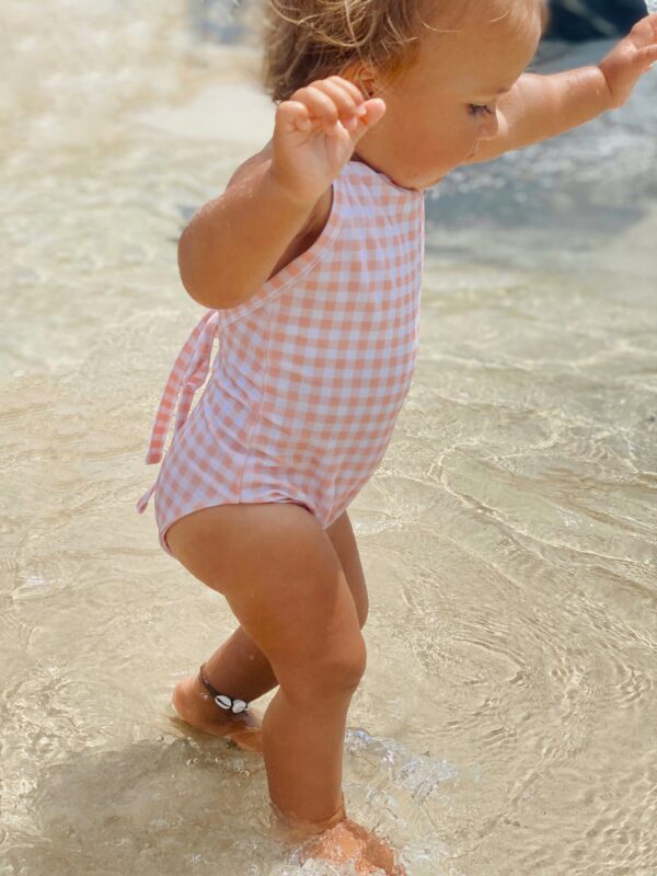A baby girl in a Mara One-Piece - Apricot Gingham swimsuit standing in the water.