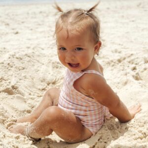 A baby girl sitting in the Mara One-Piece - Apricot Gingham on the beach.
