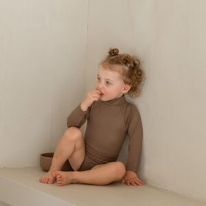 A little girl sitting on a shelf in an Essentials Range - June One-Piece - Tort Colour bodysuit.
