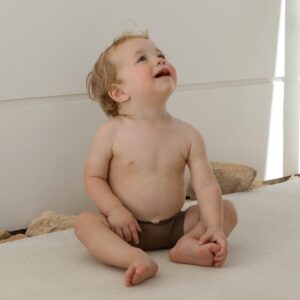 A baby sitting on the floor in a Essentials Range - Lumi Short Swim Nappy - Tort Colour diaper.