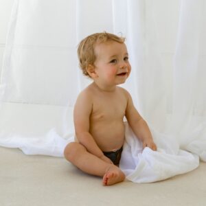 A baby sitting on the floor in front of a white curtain, wearing the Essentials Range - Lumi Short Swim Nappy in Tort Colour.