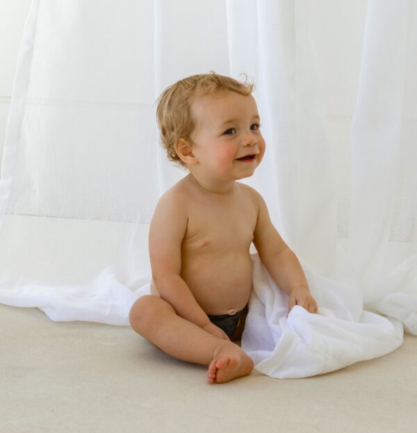 A baby sitting on the floor in front of a white curtain, wearing the Essentials Range - Lumi Short Swim Nappy in Tort Colour.
