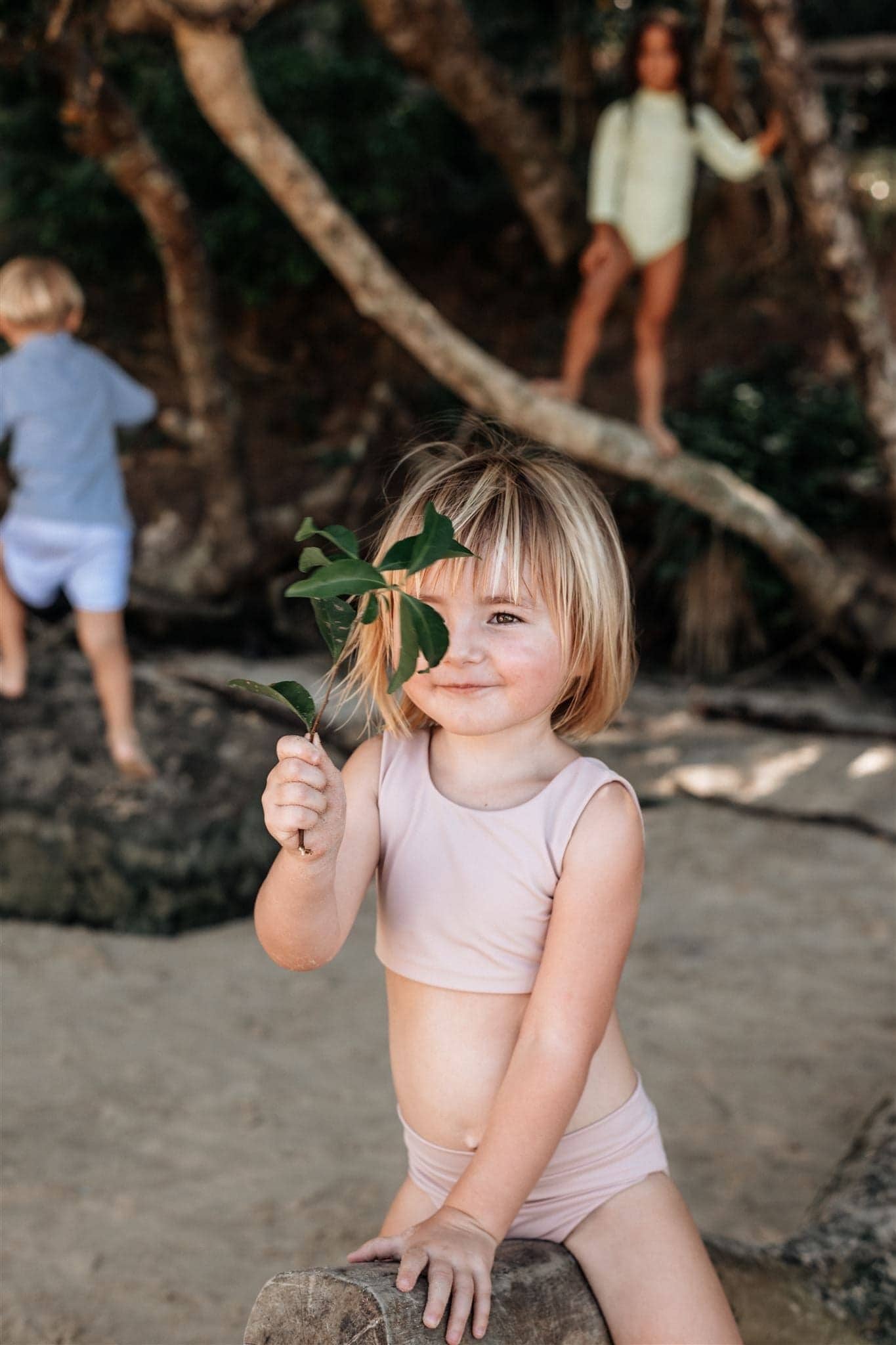 A little girl wearing sustainable swimwear holds a leaf on the beach.