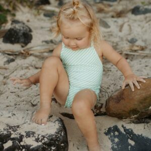 A little girl in a Retro Wave By Ina - Mara One-Piece - Fern Stripe swimsuit sitting on rocks.