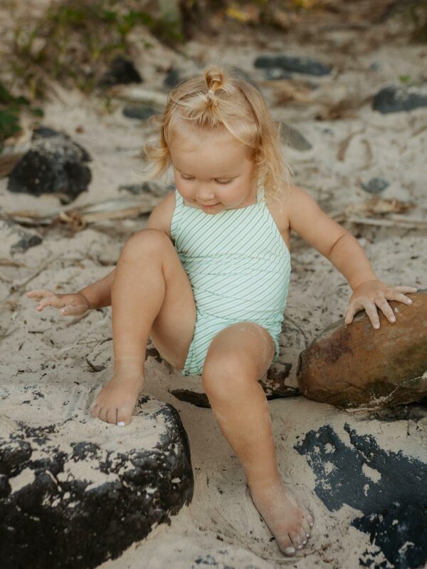 A little girl in a Retro Wave By Ina - Mara One-Piece - Fern Stripe swimsuit sitting on rocks.